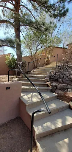 Concrete outdoor stairs with a curved black handrail leading up toward a tan building wall under trees.