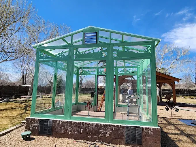 Green glass pavilion structure in a park, with people inside and a wooden gazebo nearby.