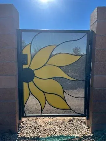 A black metal pedestrian gate between stone pillars, featuring a decorative, yellow-petaled sunflower design.