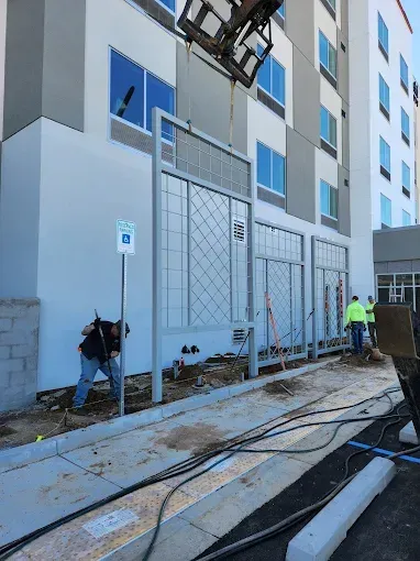 Workers installing a fence beside a white apartment building, with cables and construction materials on the ground.