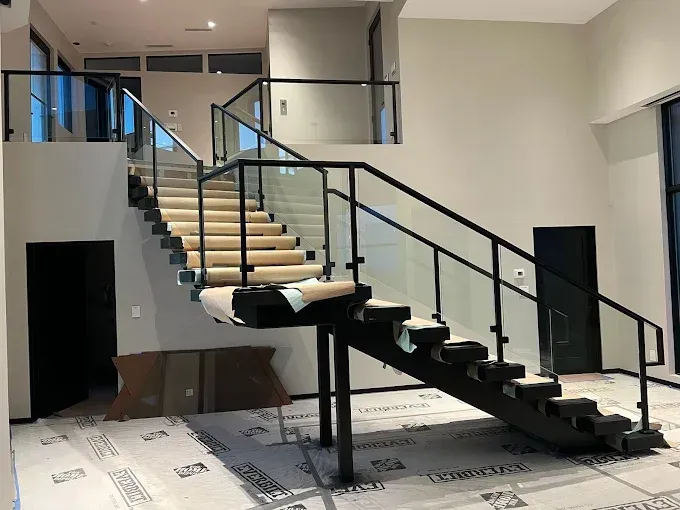 Modern staircase with black metal frames, glass railings, and light-colored treads in an unfinished construction space.