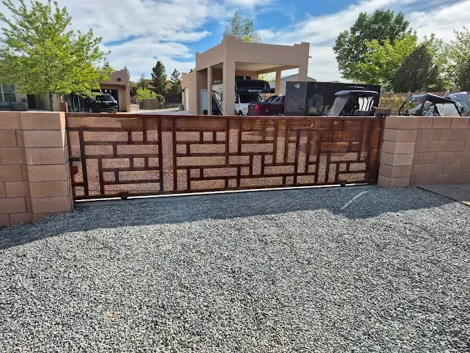 A brown, decorative metal driveway gate with a geometric pattern sits between two tan concrete block walls.