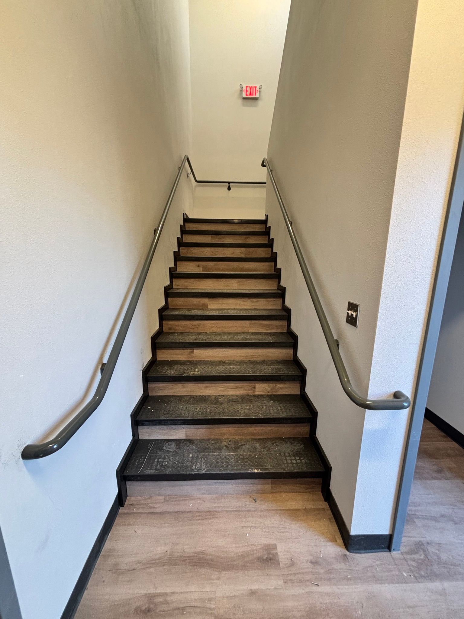 Narrow indoor staircase with metal handrails and a red exit sign at the top.