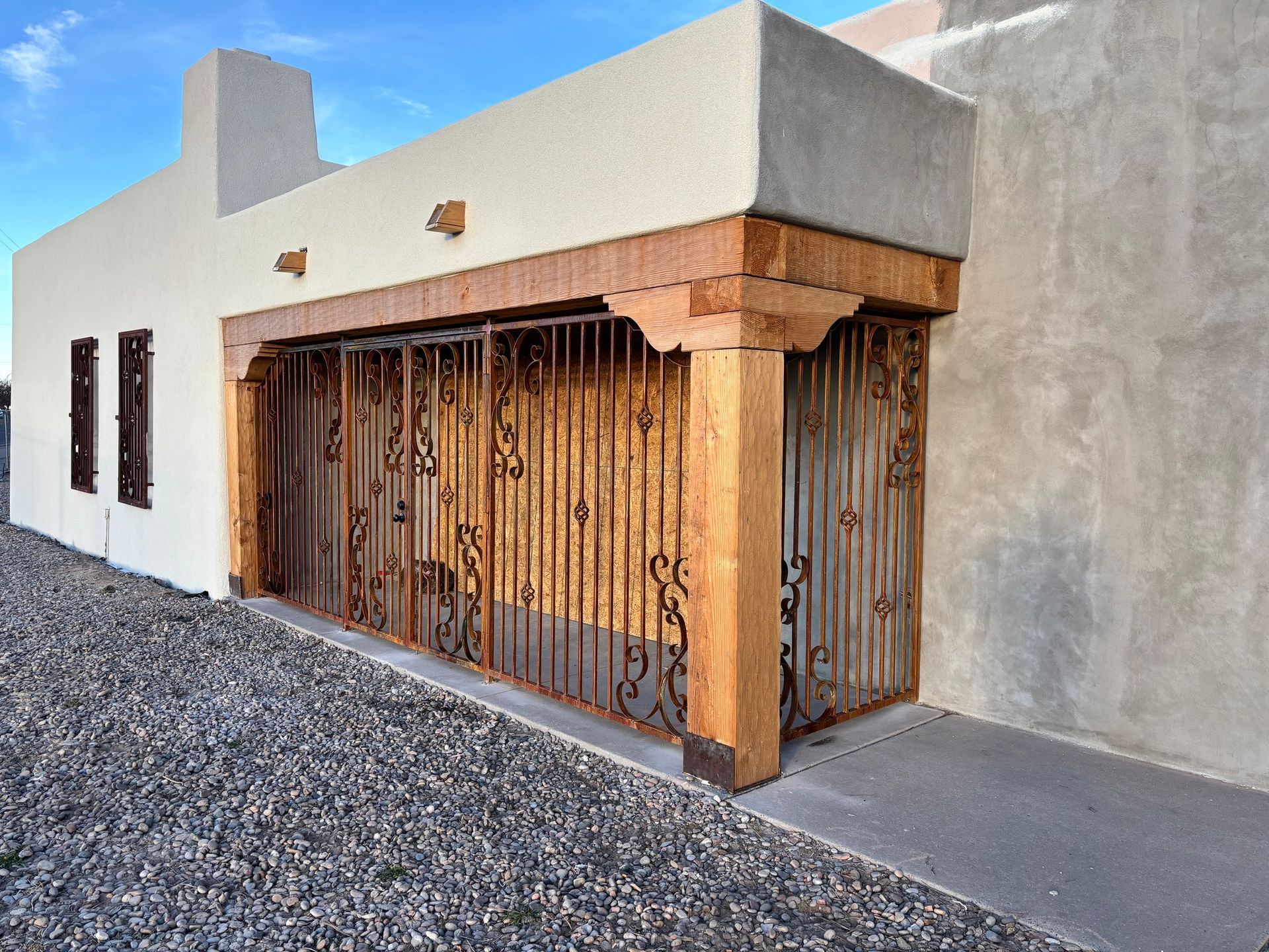Adobe-style house exterior with stucco walls, wooden porch posts, and a metal gate by gravel ground