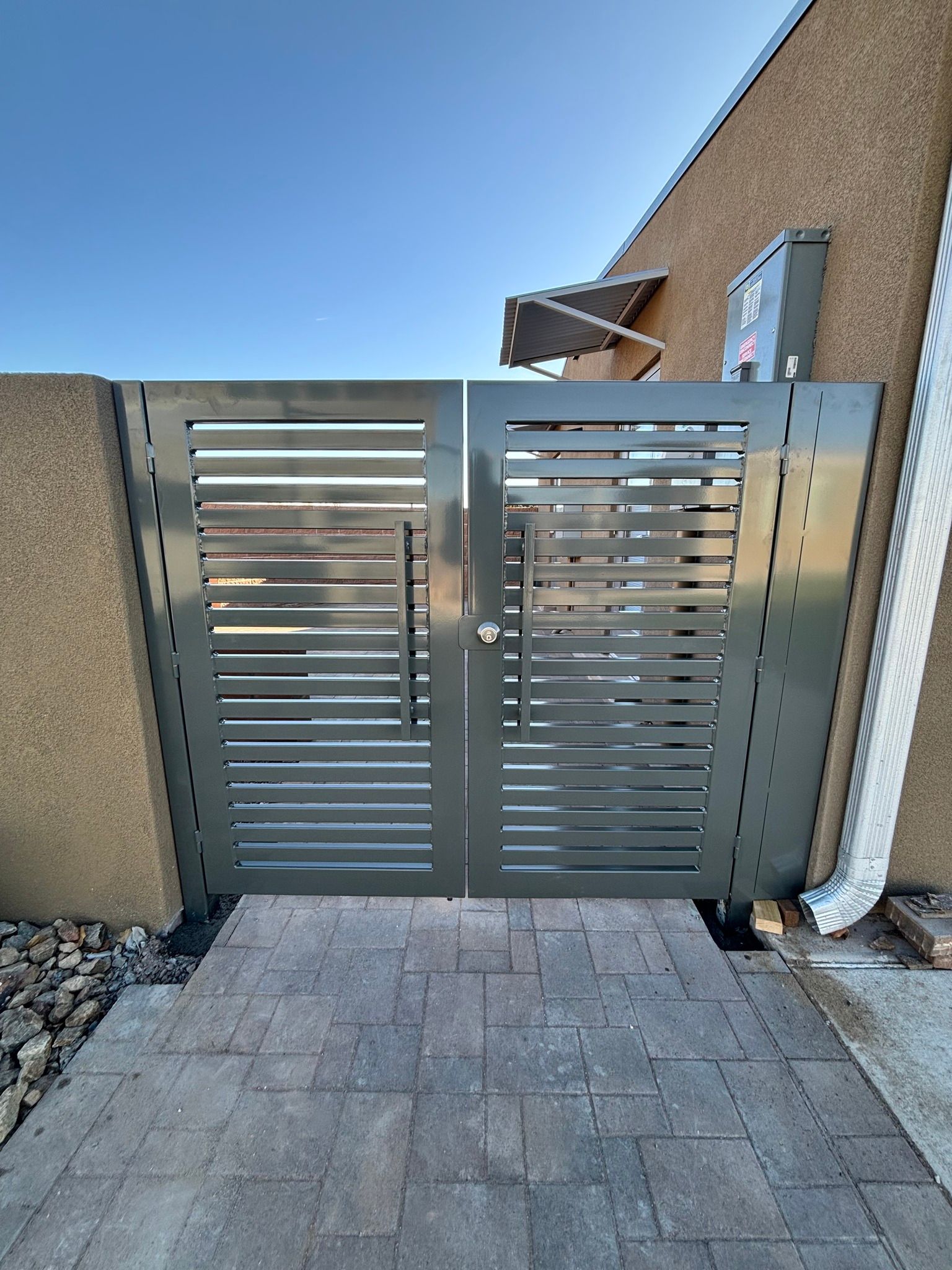 Silver metal gate at the end of a paved walkway beside a stucco house under a clear blue sky