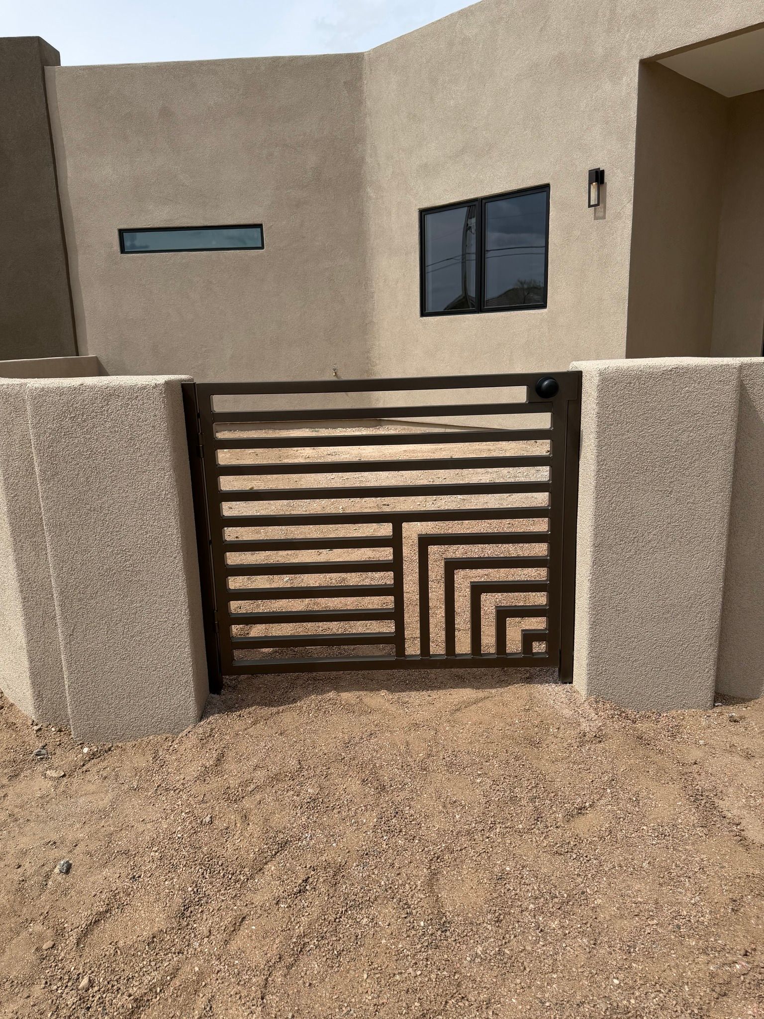 A dark metal gate with horizontal slats and a geometric corner design between two stucco walls in a desert yard.