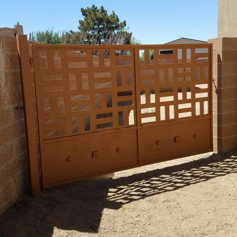 Brown metal driveway gate with geometric cutouts, set between stucco walls in bright sunlight.