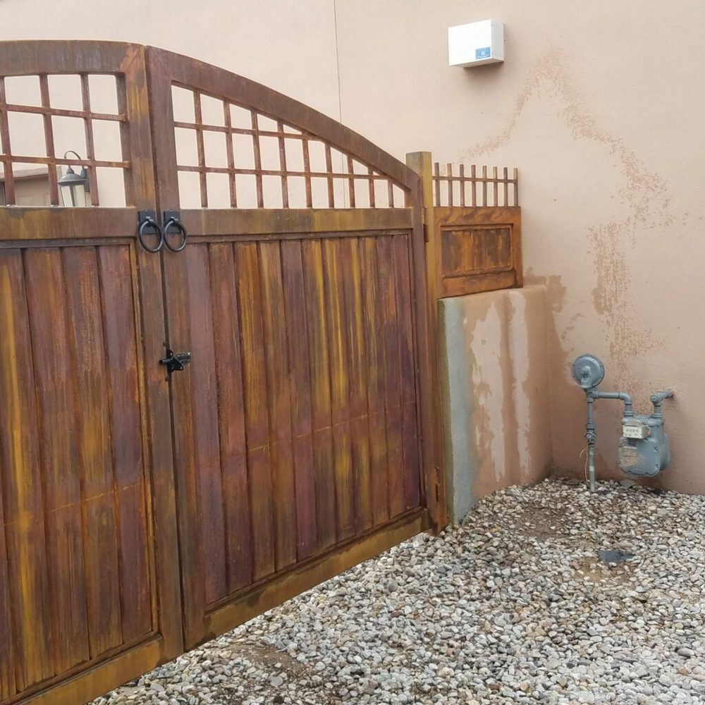 Wooden gate beside a beige stucco wall with utility pipes and gravel ground.