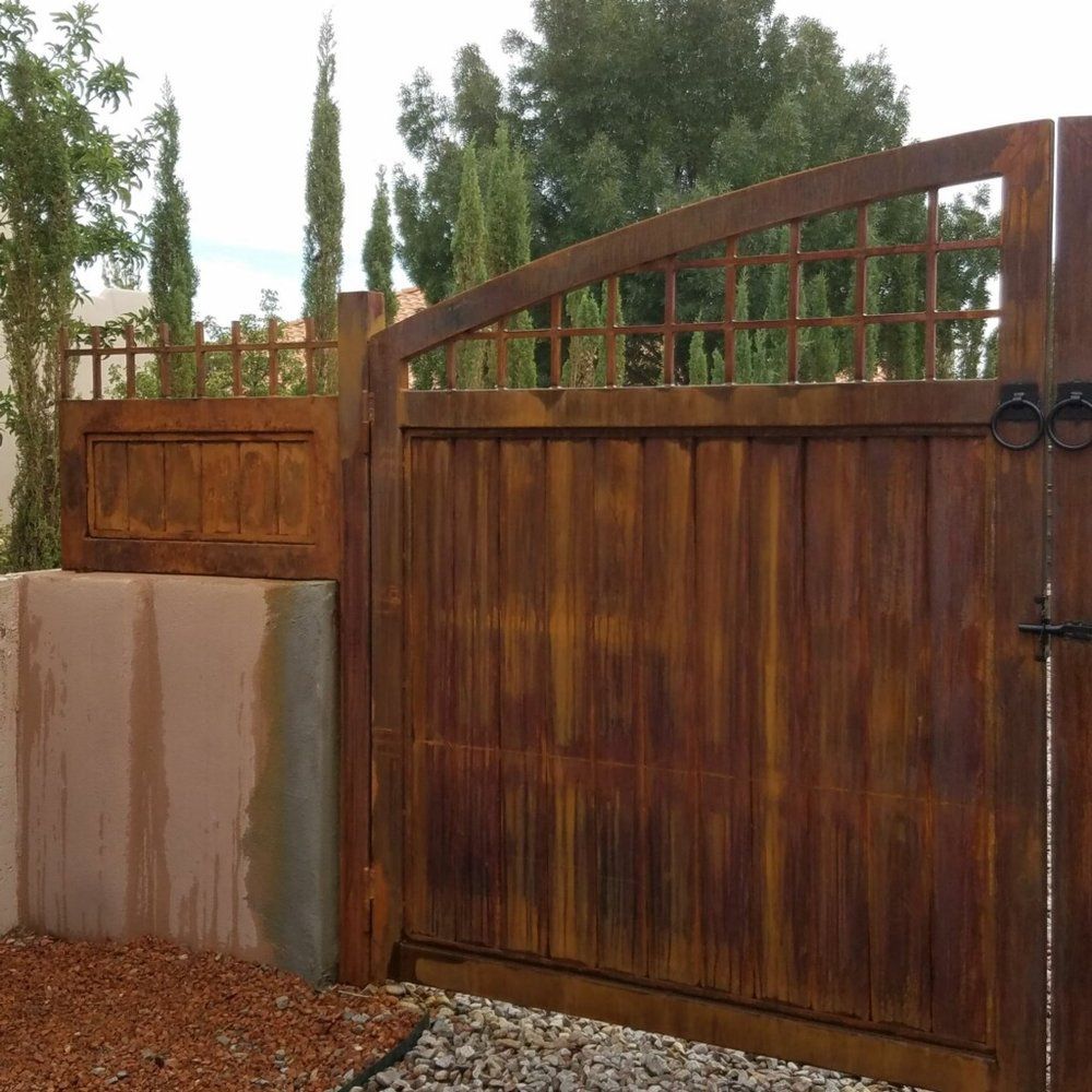 Wooden driveway gate with concrete wall, reddish gravel, and trees behind it