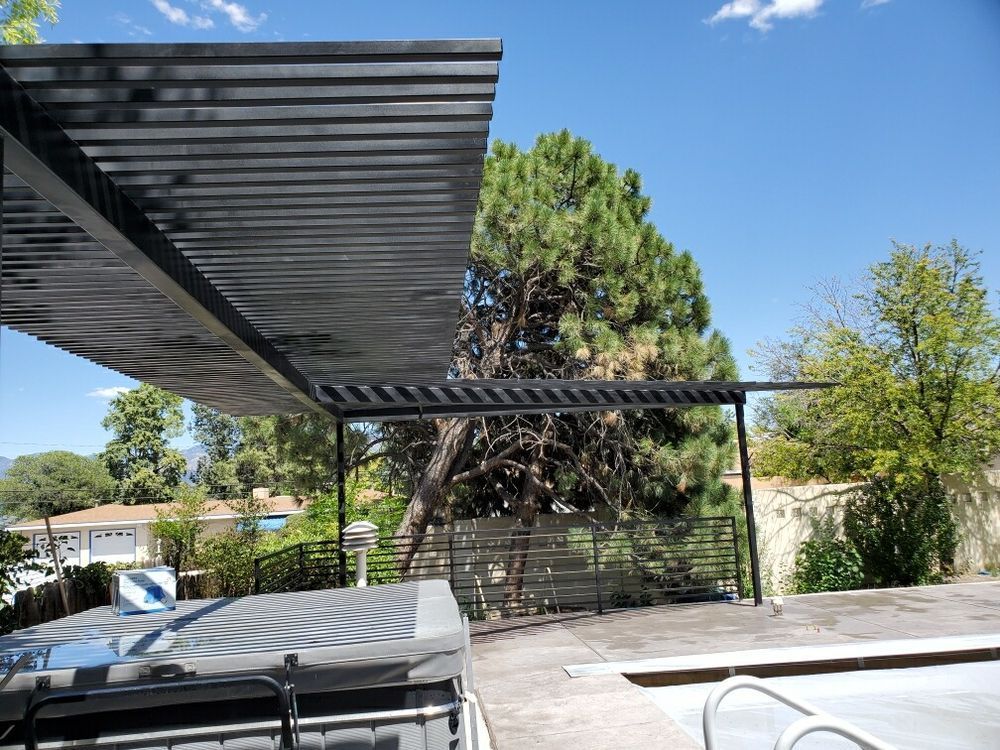 A black louvered patio cover shades a hot tub next to a swimming pool on a sunny day with trees in the background.