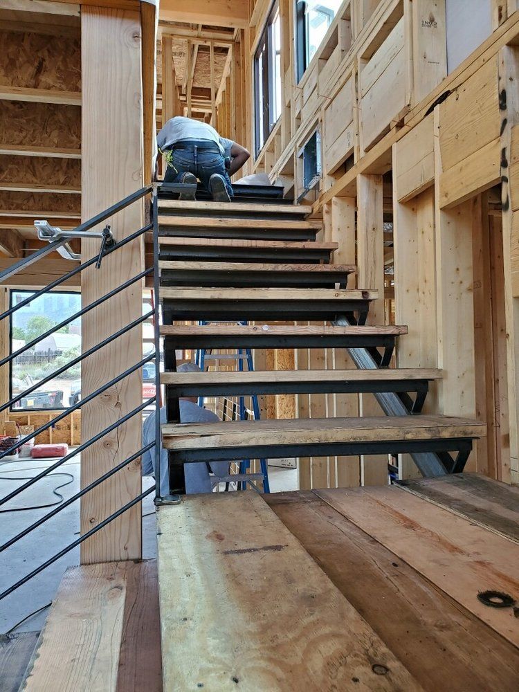 A worker kneels on wooden stairs under construction in a wood-framed building with a metal railing.
