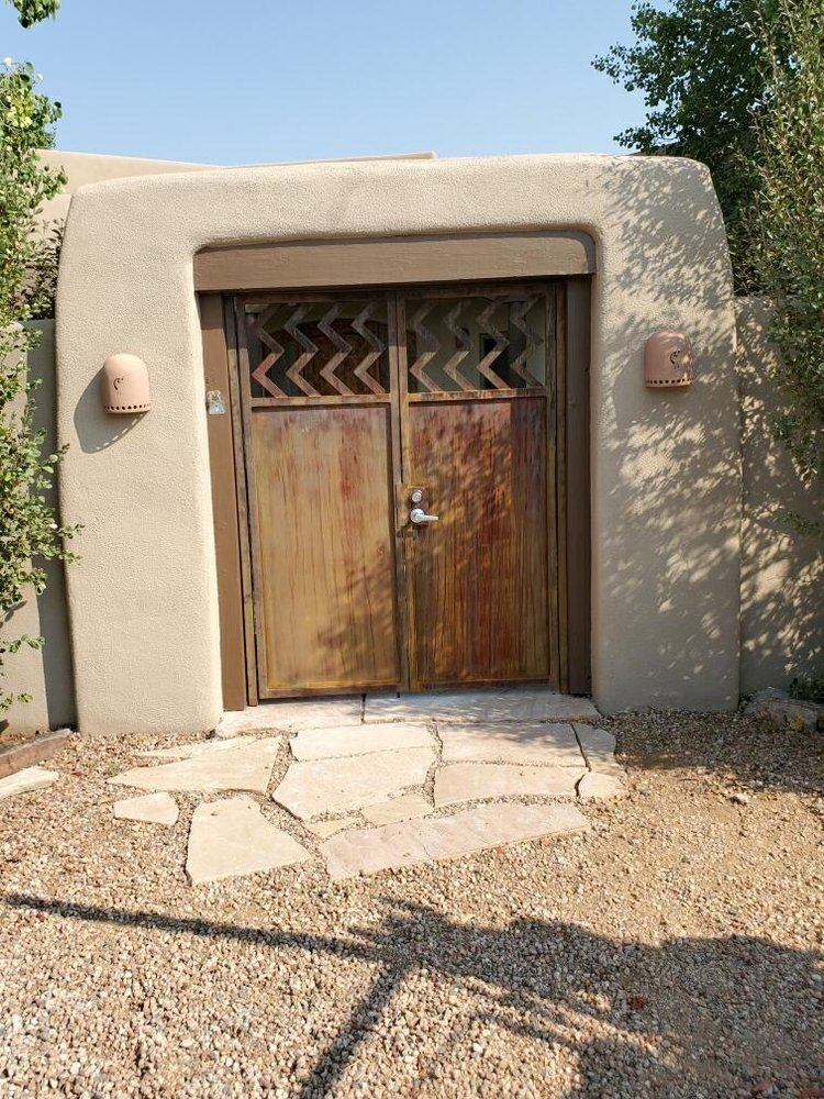 A wooden double door with a zigzag decorative top, set into a tan stucco archway surrounded by desert landscaping.