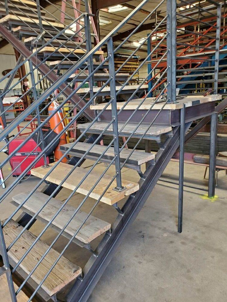 Staircase in a workshop with raw steel framing, horizontal railings, and wooden treads under construction.