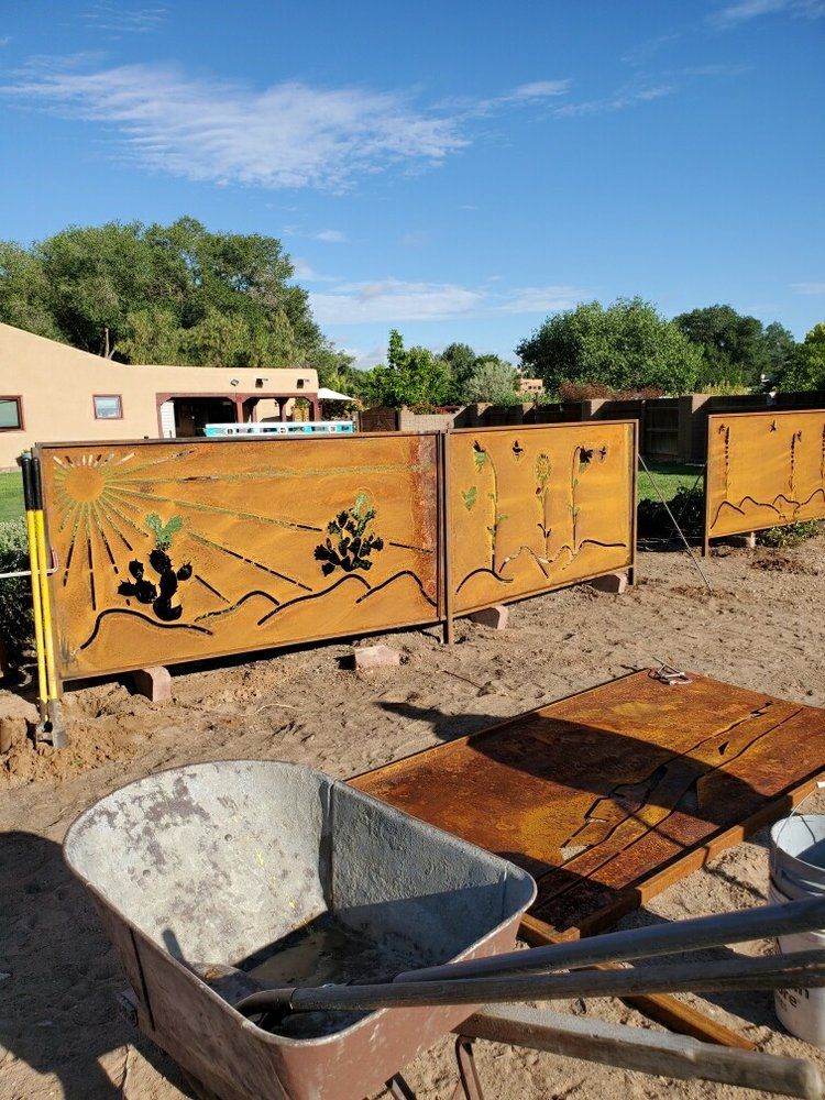 Decorative rusty metal fencing panels with cactus designs installed in a sandy backyard near a wheelbarrow.