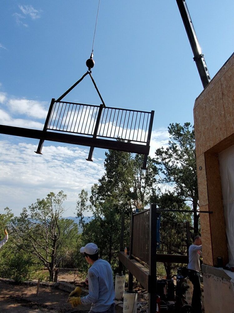 A crane hoists a metal balcony structure toward an unfinished building while two workers prepare to guide it into place.