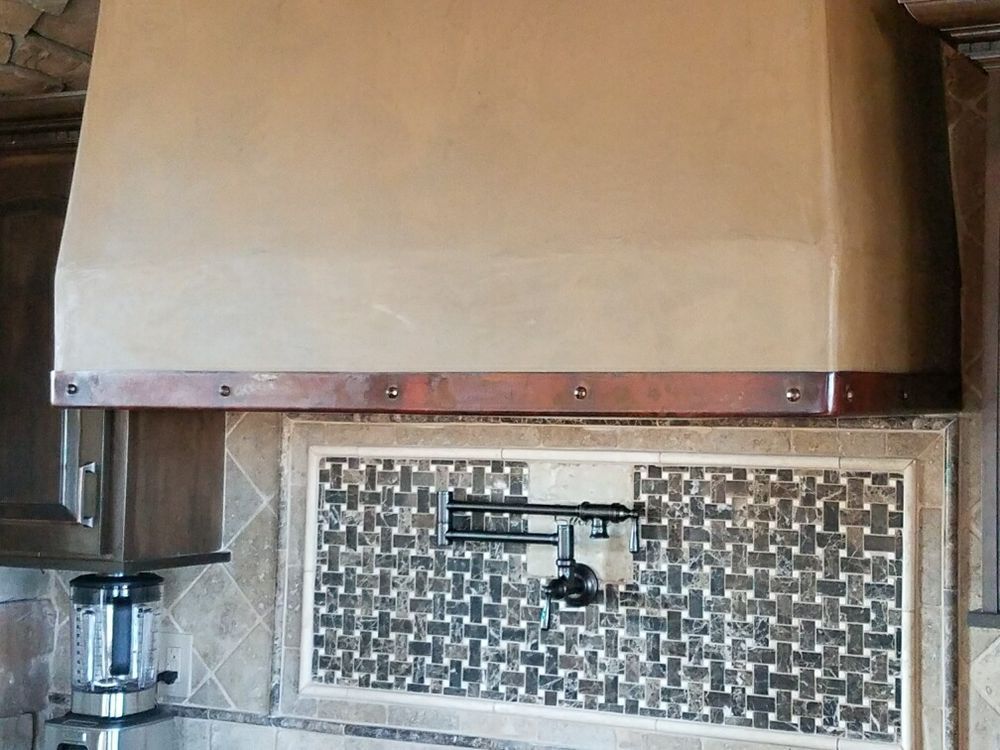 A large, light-brown plaster vent hood with a copper metal trim band above a tiled backsplash with a mounted pot filler.