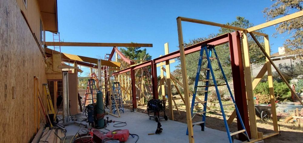 Construction site showing a house addition with exposed wood framing and red steel support beams under a clear blue sky.