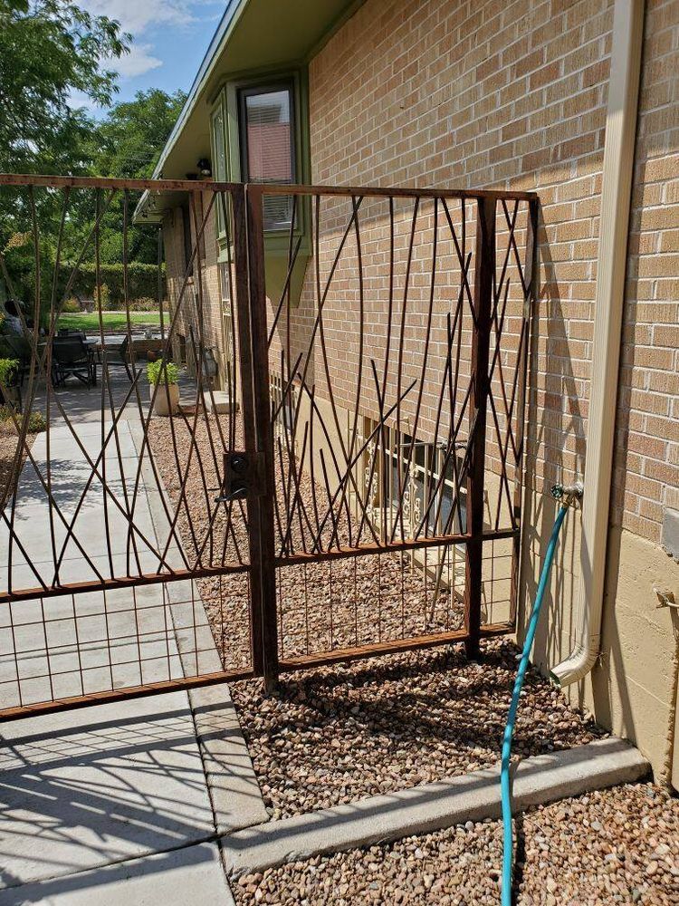 A metal gate with a twig-like design attached to the side of a brick house with a gravel ground and a blue garden hose.