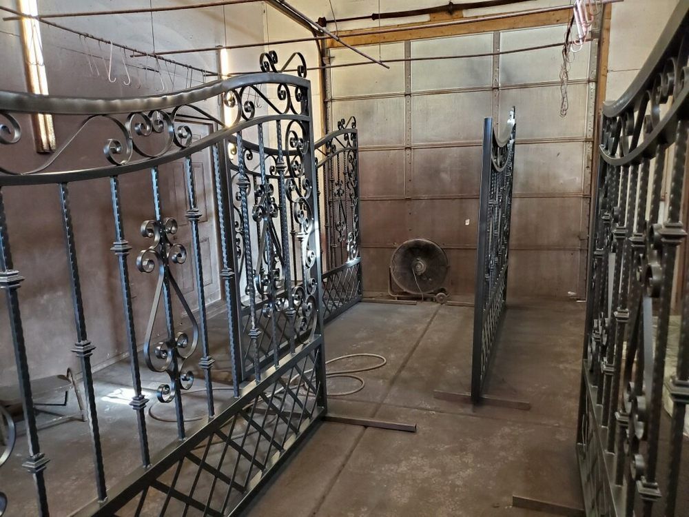 Large, dark metal wrought iron gates stand upright in a spray booth, ready for coating.