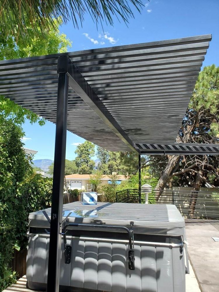 A grey hot tub under a black, slanted metal roof structure in a sunny backyard setting with trees and houses nearby.