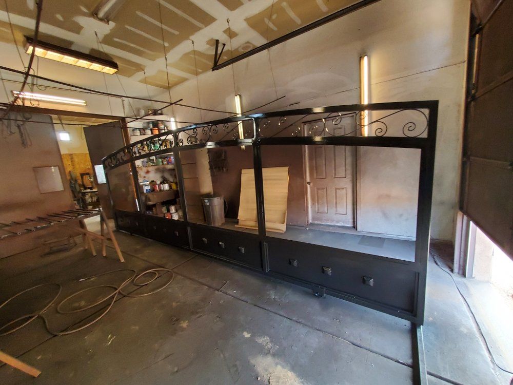 A long, arched black metal frame structure stands in a workshop setting, featuring decorative ironwork at the top.