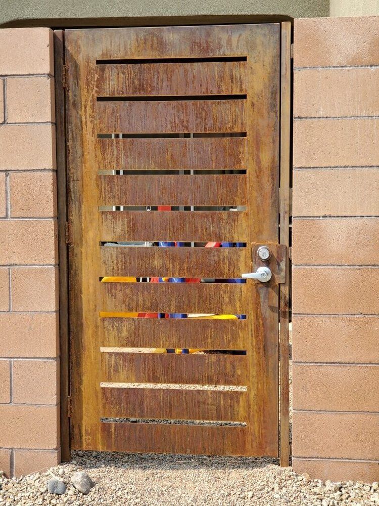 A rusted metal gate with horizontal cutouts revealing colorful glass inserts, set between two tan concrete block walls.