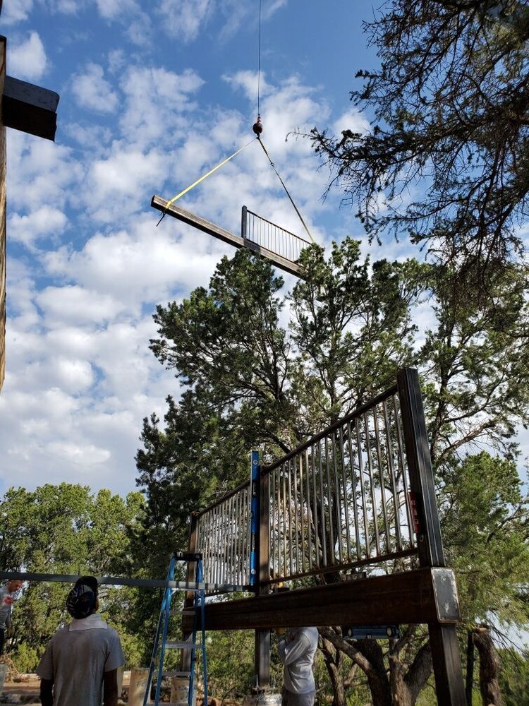 A crane lifts a large metal staircase section through trees toward an outdoor balcony under a blue, cloudy sky.