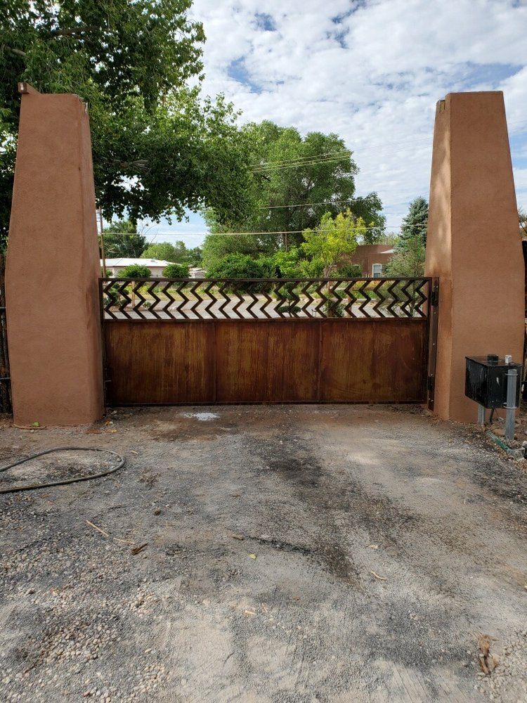 A wooden gate with a decorative chevron metal top sits between two tall, tan stucco pillars under a cloudy blue sky.