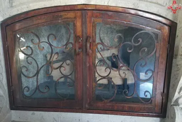 Arched fireplace doors with a dark, weathered wood frame and ornate, black iron scrollwork overlays on glass panels.