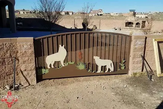 A brown metal gate featuring silhouettes of two wolves, set between tan concrete block walls in an outdoor desert area.