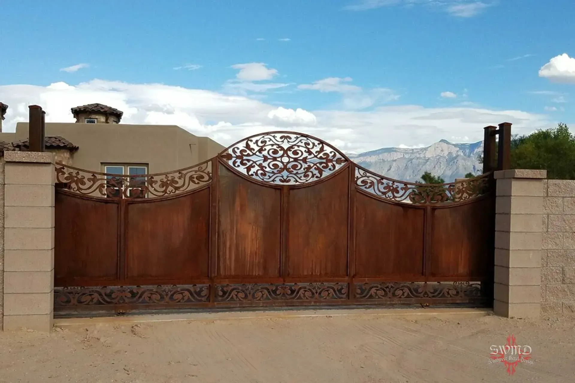 A decorative, rusted metal driveway gate with ornate scrollwork, set between two concrete pillars under a cloudy sky.