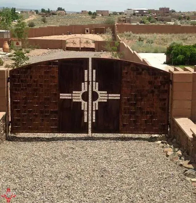 A metal double gate with a rustic brown finish and a Zia symbol cut-out design, situated on a gravel driveway.