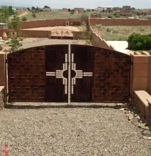 A decorative, rustic metal gate featuring a central Zia symbol, situated in a gravel driveway with a desert background.