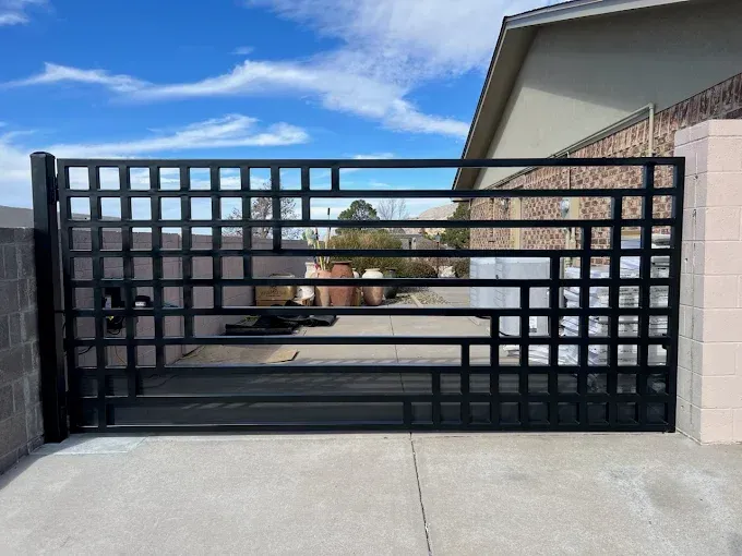 Black metal driveway gate with square grid design in front of a house, closed across a concrete driveway