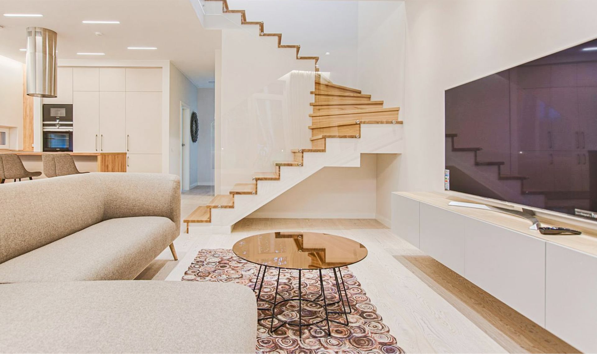 Modern living room with a light-colored couch, round glass coffee table, and wooden staircase leading to an upper level, showcasing a clean and inviting home environment ideal for deep cleaning services.