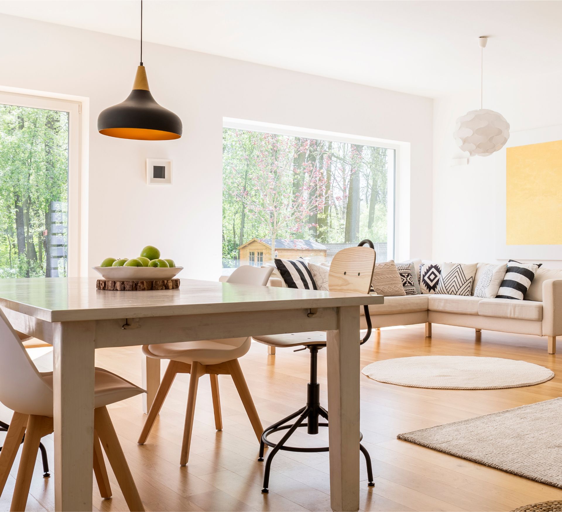 Bright and modern living space featuring a wooden dining table with a bowl of green apples, stylish chairs, a cozy sofa with decorative pillows, and large windows showcasing a lush outdoor view, emphasizing the benefits of a clean and inviting home.