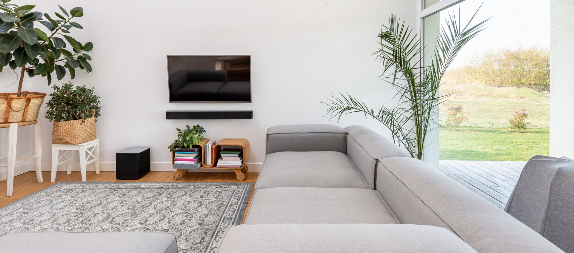 Modern living room with a gray sectional sofa, wall-mounted television, decorative plants, and a cozy atmosphere, emphasizing cleanliness and comfort for recurring house cleaning services.