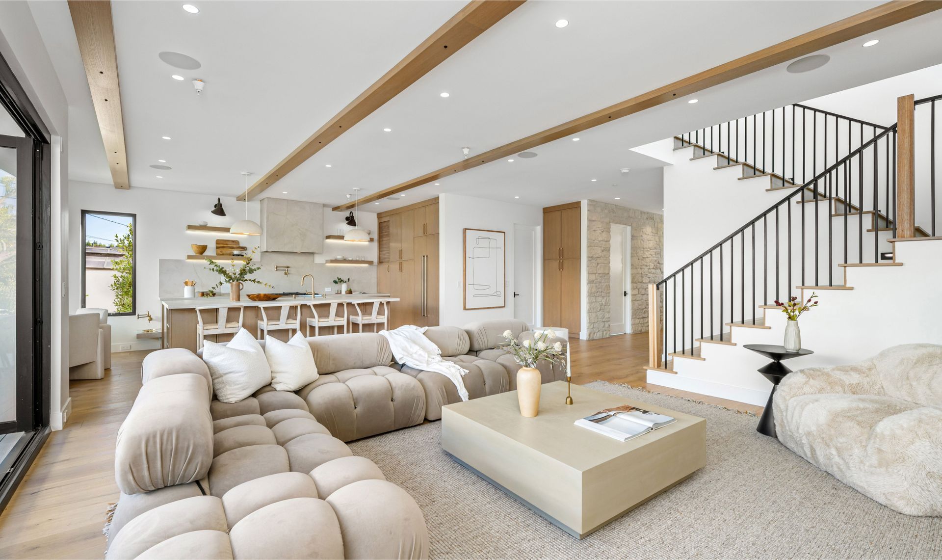 Modern living room with a beige sectional sofa, light wood coffee table, and a stylish kitchen in the background, showcasing a clean and inviting space ideal for recurring house cleaning services.