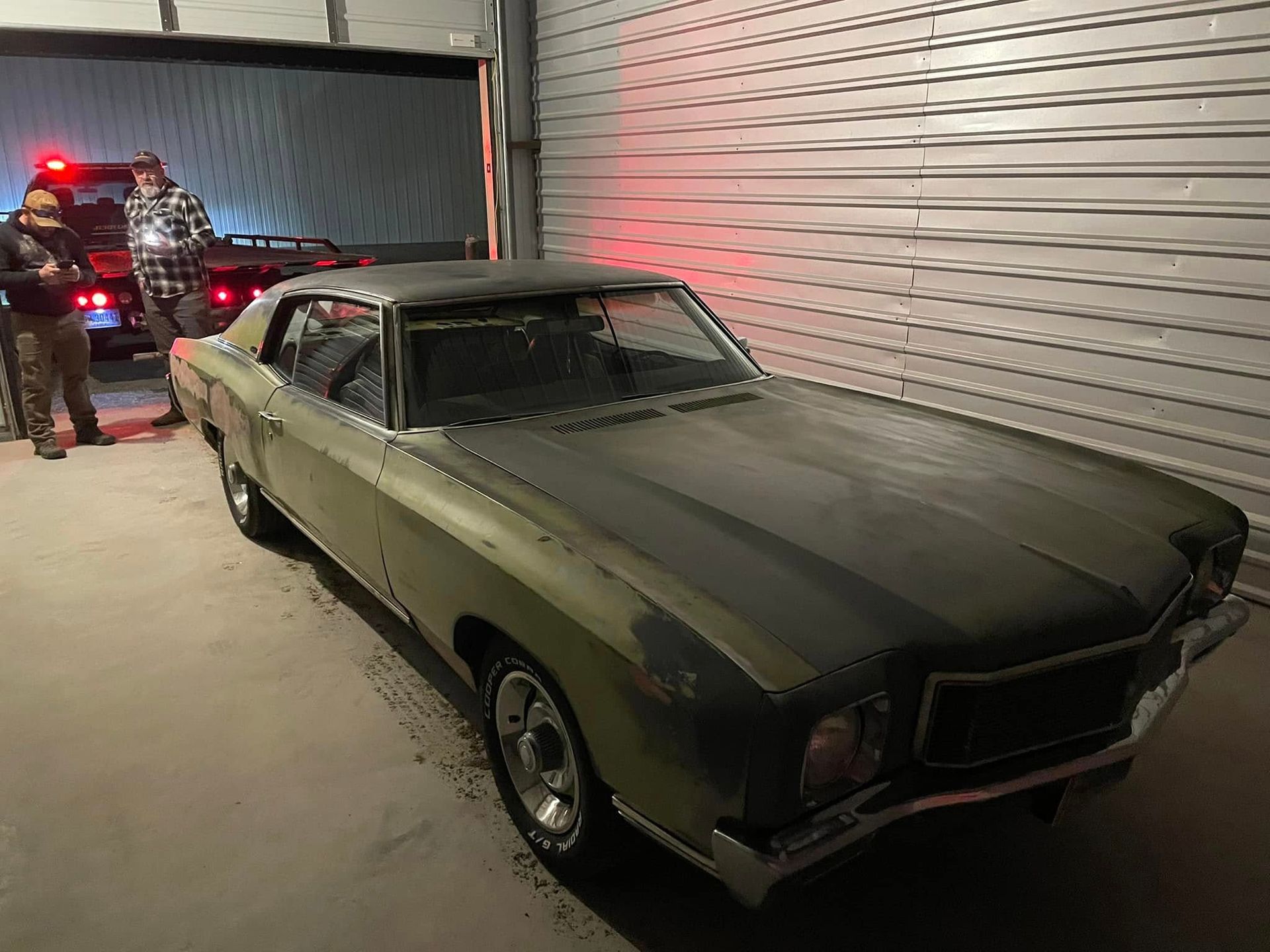 A green and black classic car is parked in a garage with two people standing beside it; the garage door is slightly open.