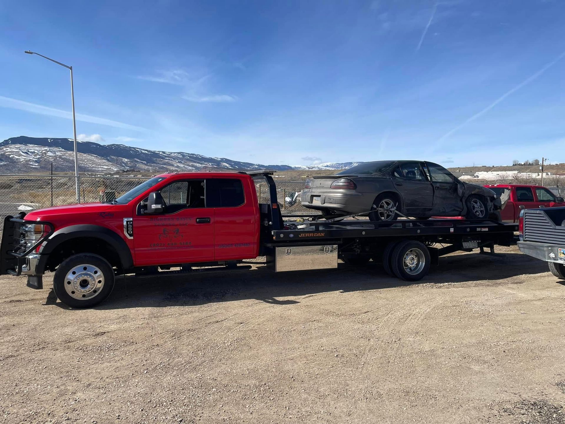 A red tow truck carrying a damaged gray car on a flatbed, outdoors in a sunny setting with mountains in the background.