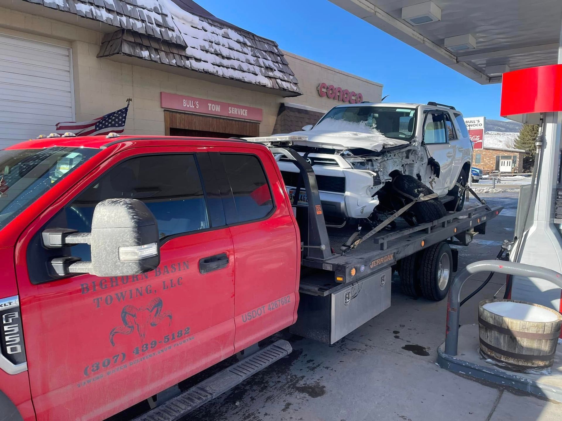 Red tow truck hauling a severely damaged white SUV at a gas station. The SUV's front end is crushed.