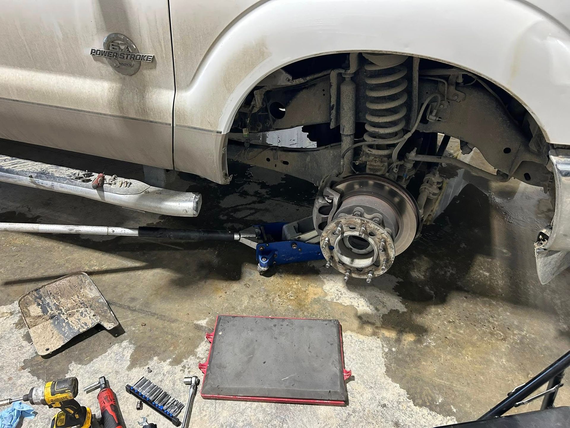 A white truck's front wheel is removed, revealing suspension components. Tools and a jack sit on a concrete garage floor.