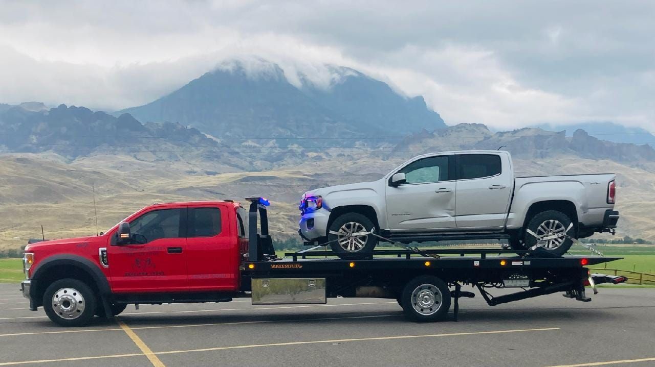 A red tow truck carrying a silver pickup truck on a flatbed, set against a mountainous landscape under a cloudy sky.