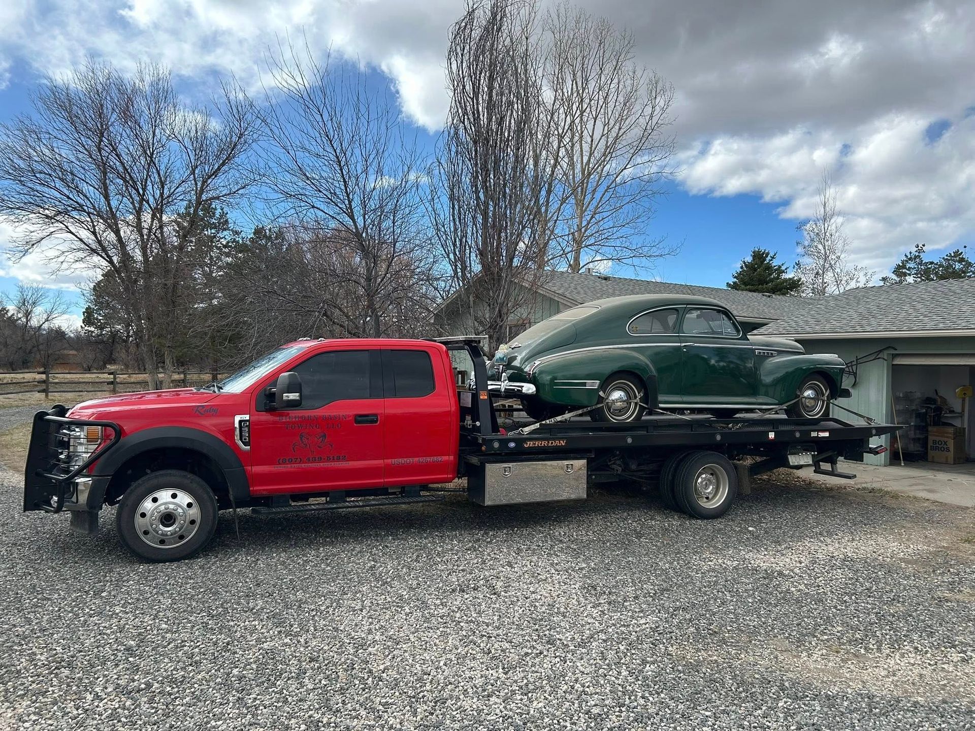 Red tow truck with a green vintage car loaded on its flatbed, parked in a gravel driveway with a house in the background.