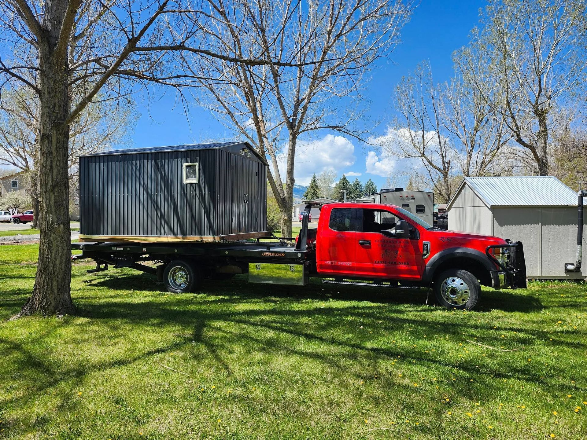 A red flatbed truck carrying a small black shed on a sunny day in a grassy area with trees and a white shed.