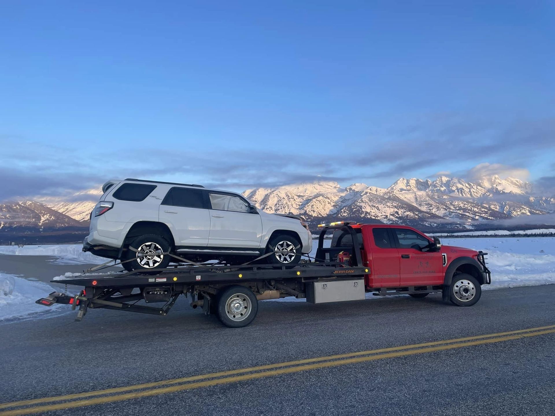 A white SUV is being towed on a red flatbed truck along a snowy road with a mountain range in the background.