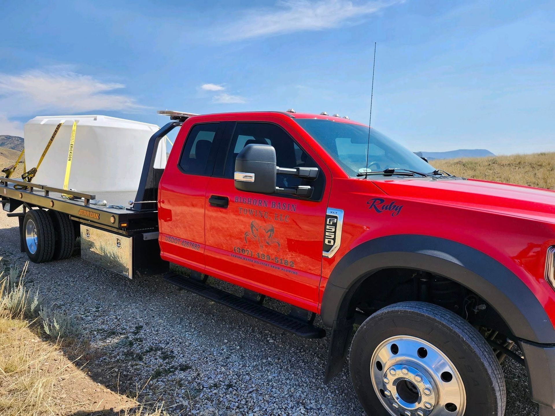 Red tow truck carrying a large white cylindrical container on its flatbed in a desert setting.