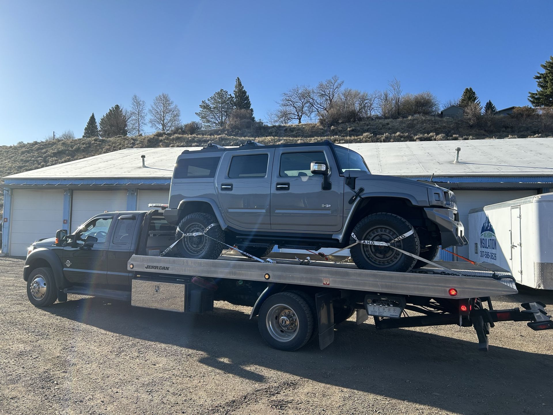 A gray Hummer SUV is secured on a flatbed tow truck. The truck is parked in front of a building under a clear sky.