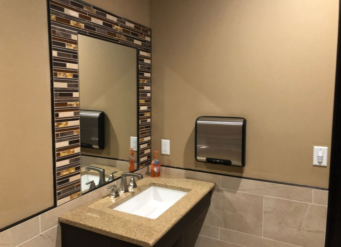 Bathroom with a sink, mirror, soap dispenser, and hand dryer. Beige walls, stone countertops, and brown tile.