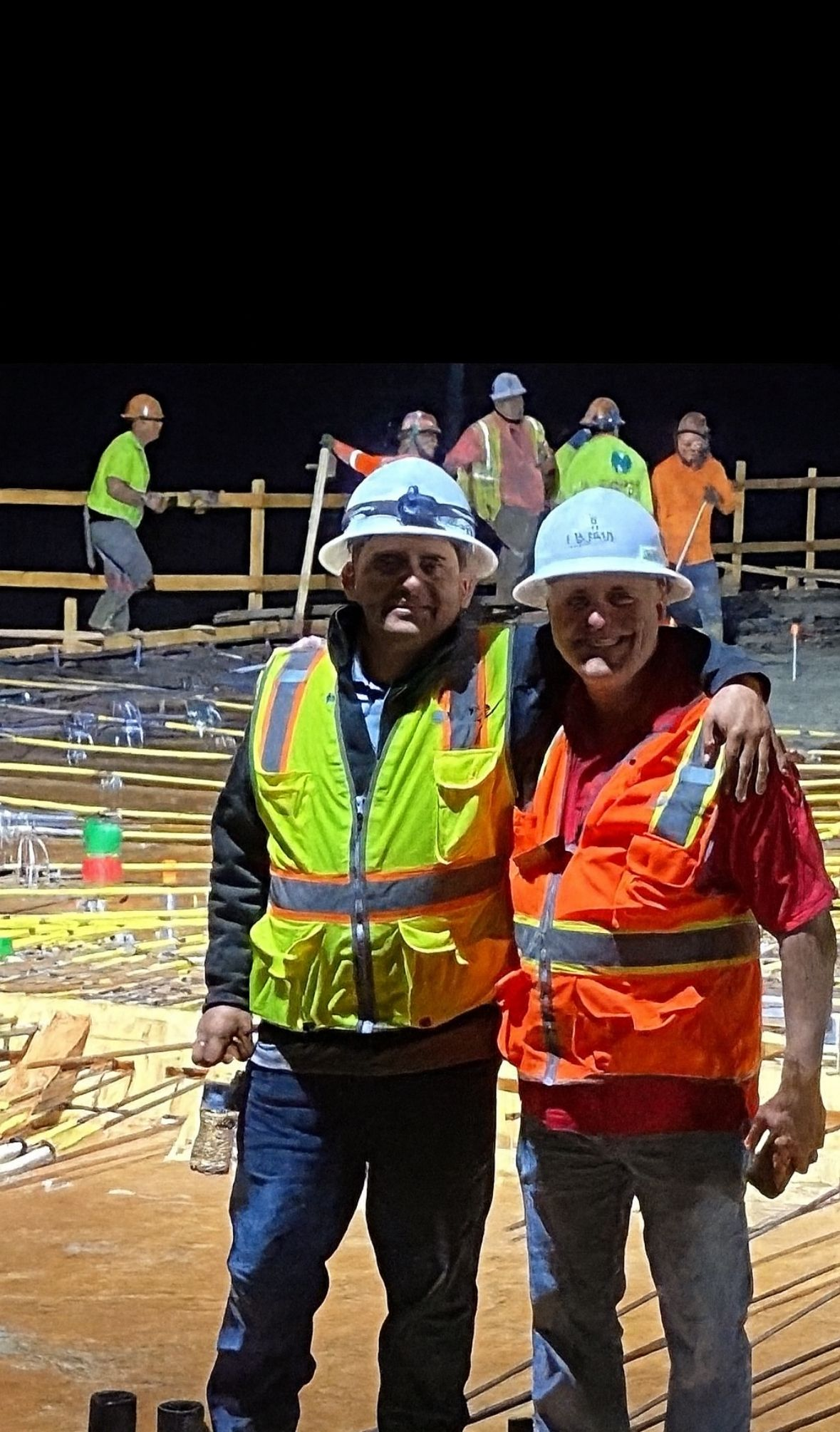 Two construction workers with arms around each other smiling, other workers in background on a construction site.